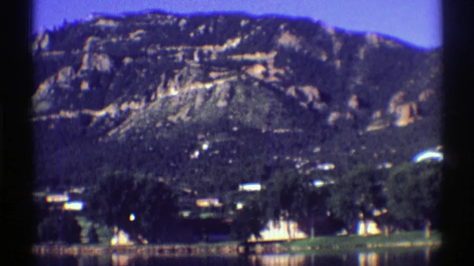 1969 Colorado 8mm frame showing a calm lake, trees, small shoreline structures, and rugged mountain slopes in the distance.