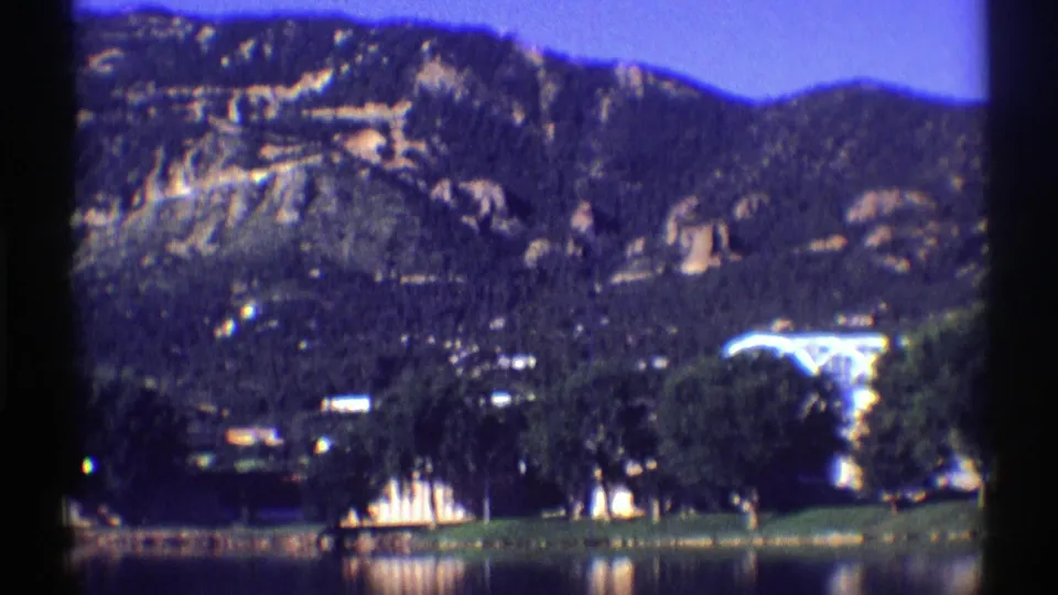 1969 Colorado lakeside landscape with a mountain ridge, dark tree cover, small shore structures, and reflective water.