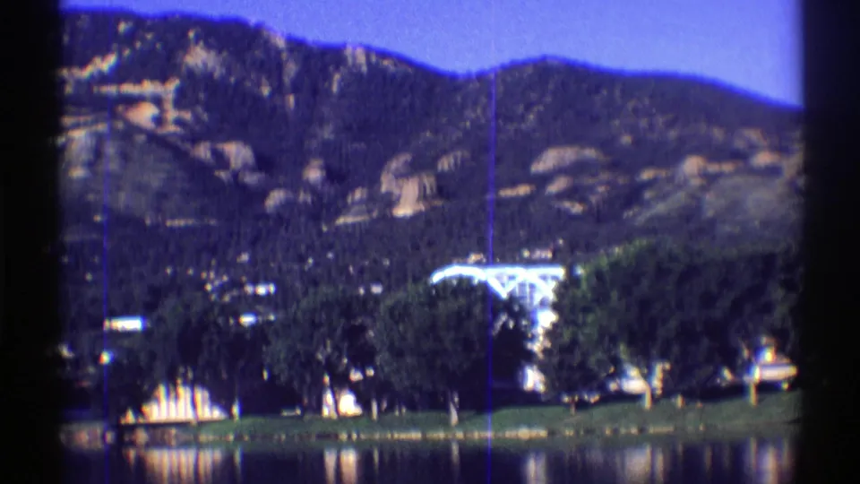 Colorado 8mm home-movie frame with lake reflections, clustered trees, a bright shoreline roofline, and rocky mountain slopes.