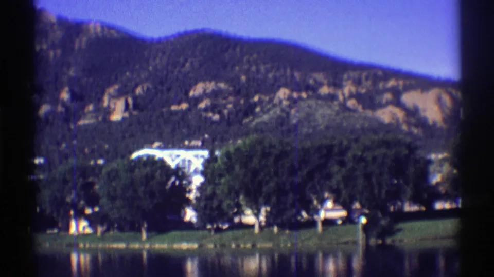 1969 Colorado landscape frame showing a quiet lake foreground, dense shore trees, a small structure, and rugged slopes beyond.