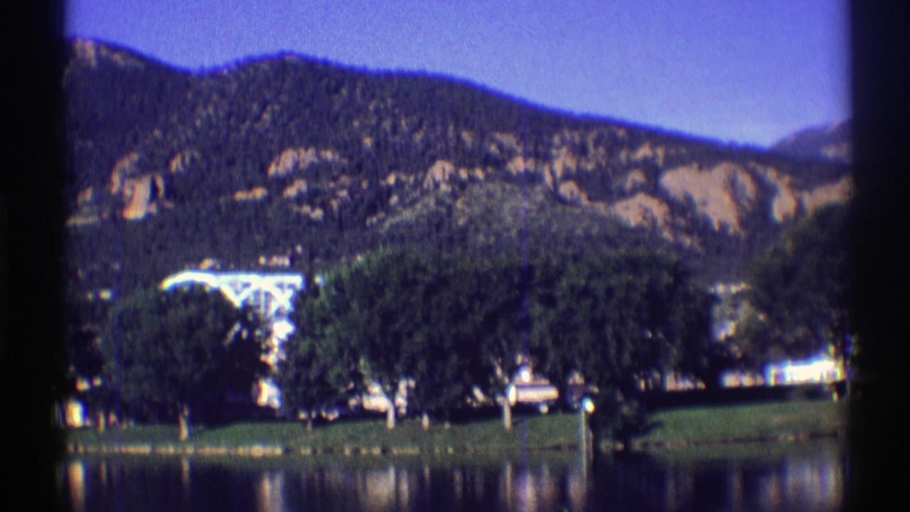 Mountain slope above trees, grass, water, and a partly hidden pale structure.