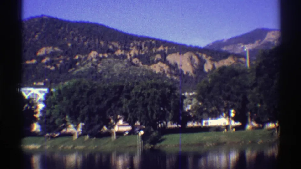 Colorado home-movie frame with mountains, a blue sky, dense green trees, water reflections, and a pale building partly obscured.