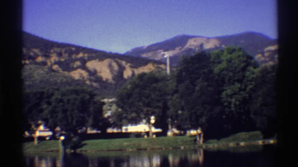 1969 Colorado landscape frame with layered mountains, lakeside trees, a pale building near the shore, and soft water reflections.