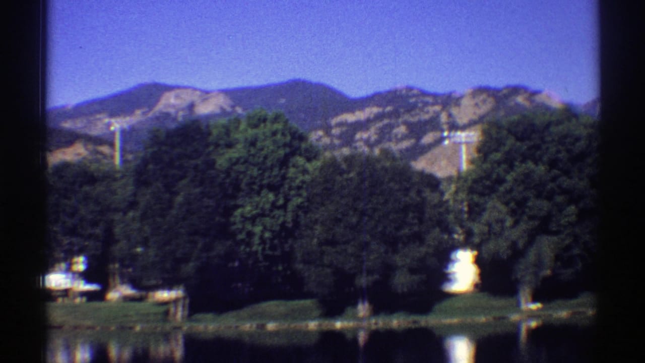 Calm lake with tree-lined shore and distant rocky mountains under a blue sky.