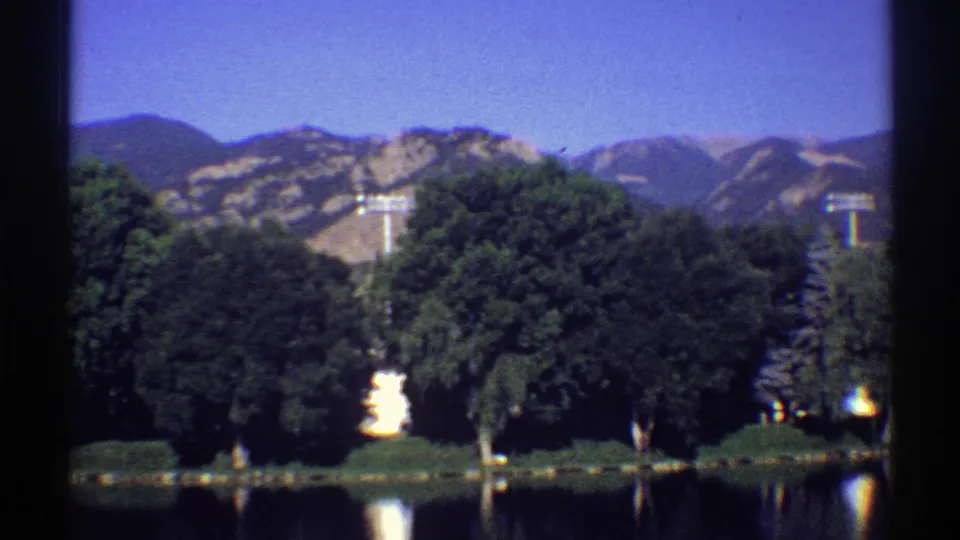 1969 Colorado 8mm frame of a wooded lakeshore, bright water reflection, and rugged mountains in the distance.