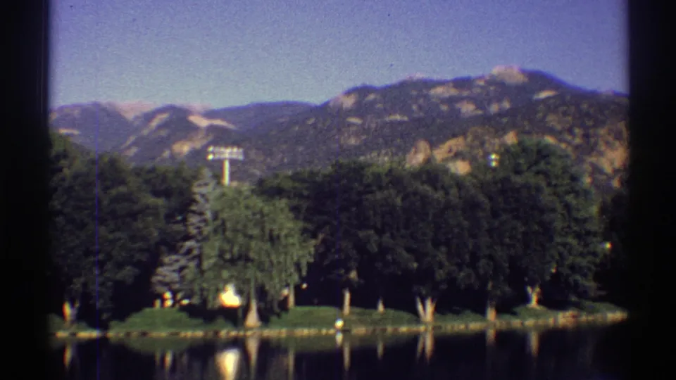 1969 Colorado 8mm frame with a broad view of lake water, mixed trees, and sunlit mountain slopes beyond the shore.