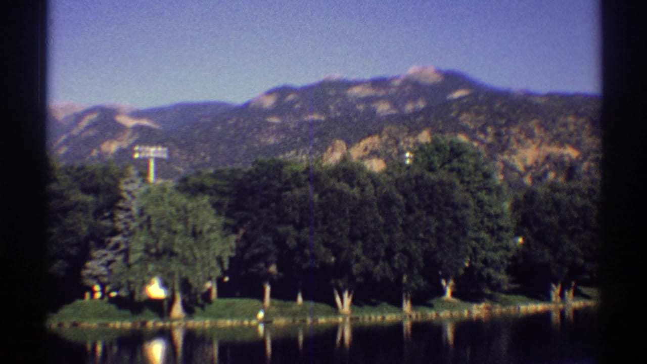 1969 Colorado 8mm frame showing a still lake, dark green shoreline trees, and rugged mountains in the background.