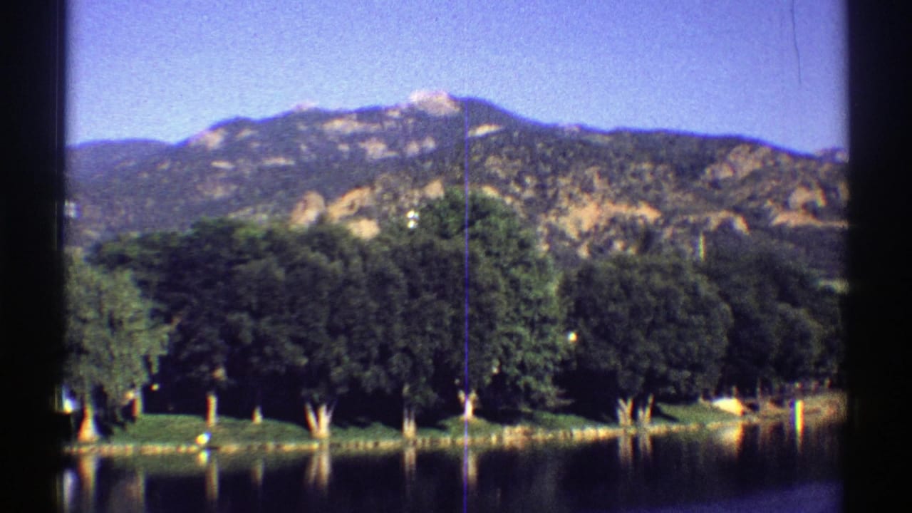 1969 Colorado 8mm view of a tree-lined lake with reflected trunks and a mountain ridge beyond the shore.