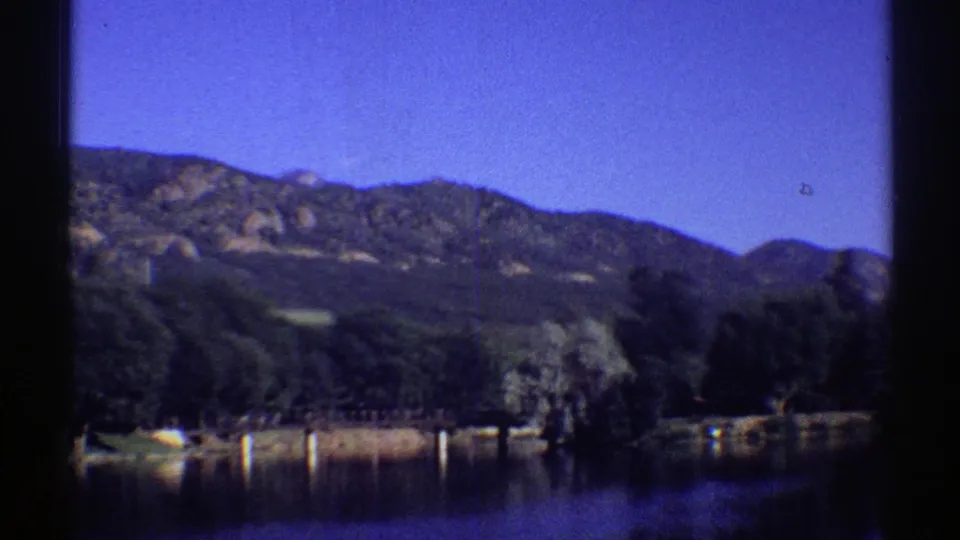 1969 Colorado 8mm frame showing dark lake water, a tree-lined shore, and distant mountain ridges under clear sky.