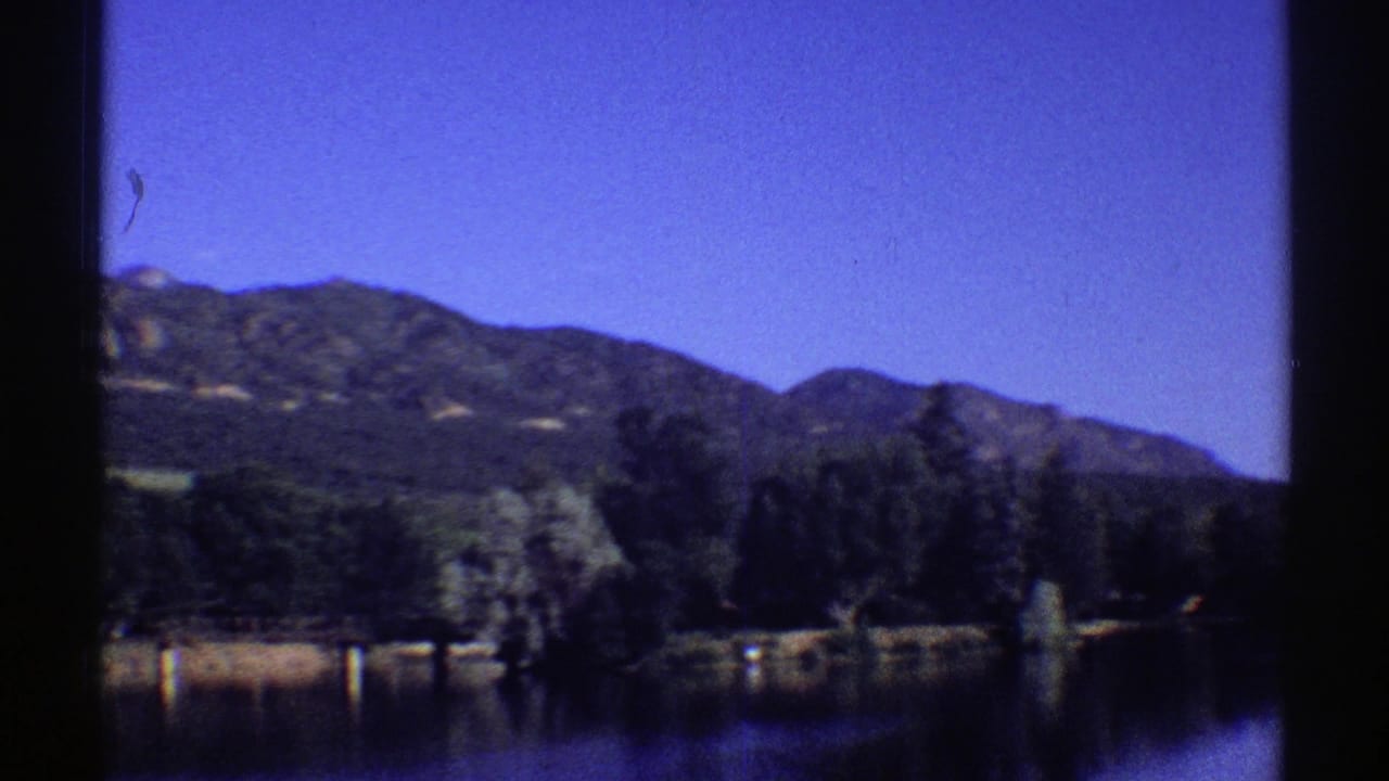 1969 Colorado 8mm frame showing a calm lake, tree-lined shore, and mountains beneath a saturated blue sky.