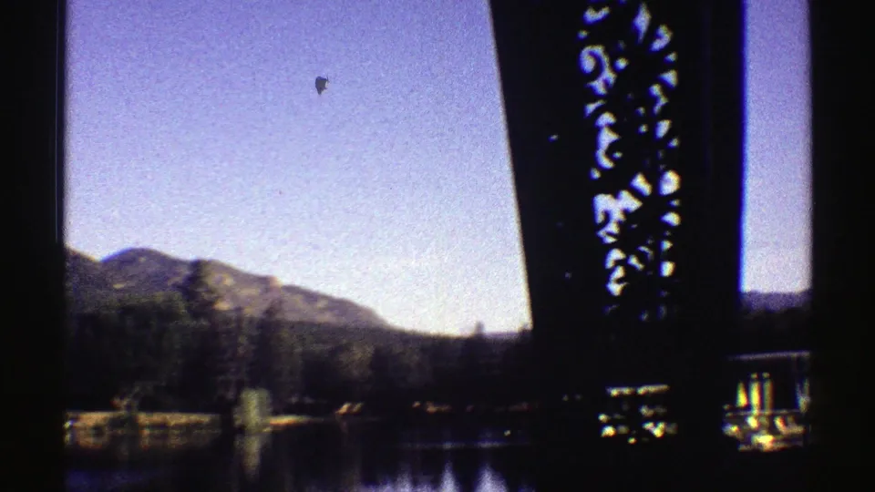 1969 Colorado lakeside view seen through dark bridge supports, with mountains, trees, and reflective water beyond.