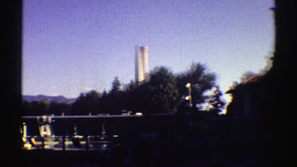 1969 Colorado lakeside marina view with boats, dock structures, trees, and a distant tall industrial smokestack.