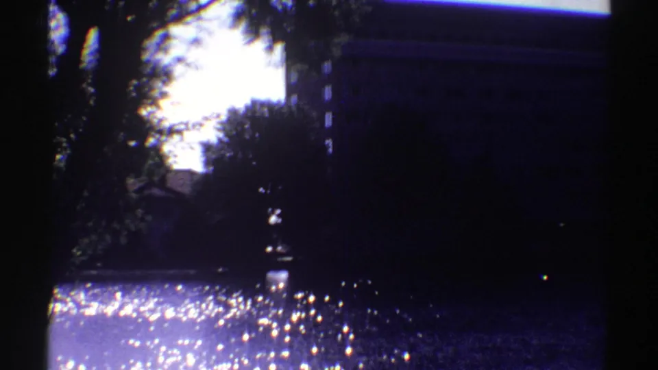 Handheld 8mm view across shimmering water, with trees in silhouette and part of a large building at the right edge.