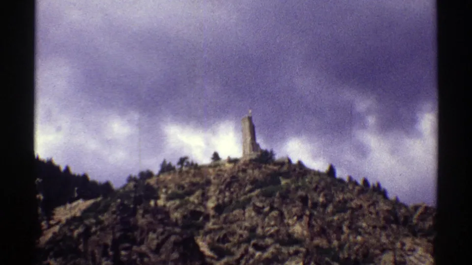 A grainy 1969 Colorado scenic view shows a wooded hill, clouded sky and a narrow structure rising from the ridge.