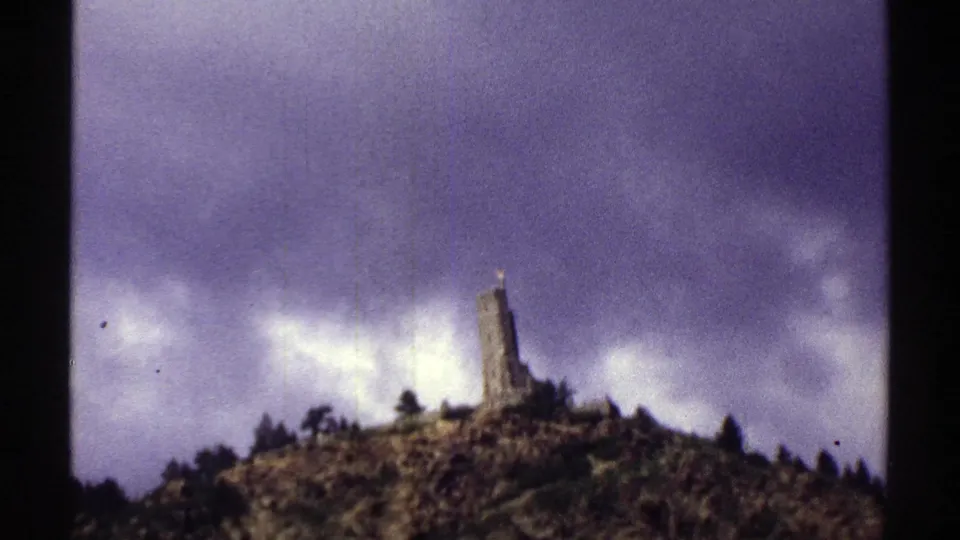 1969 Colorado 8mm view of a hilltop stone monument, sparse trees, and rough rock slope under gray-purple clouds.