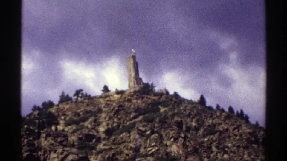 1969 Colorado 8mm frame showing the hilltop monument, rugged rocks, scattered evergreens, and moody cloud cover.