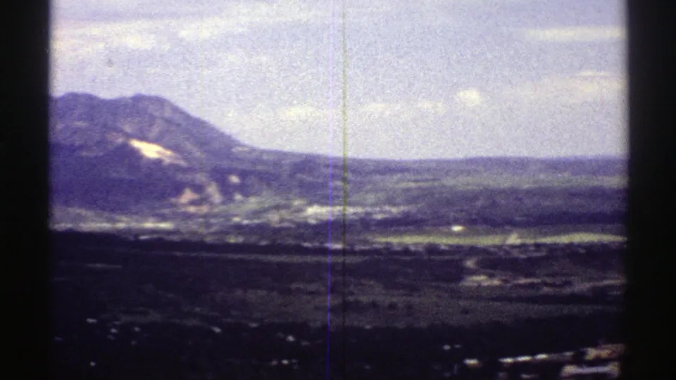 1969 Colorado home-movie frame showing a broad valley panorama with a mountain slope at left and distant structures below.