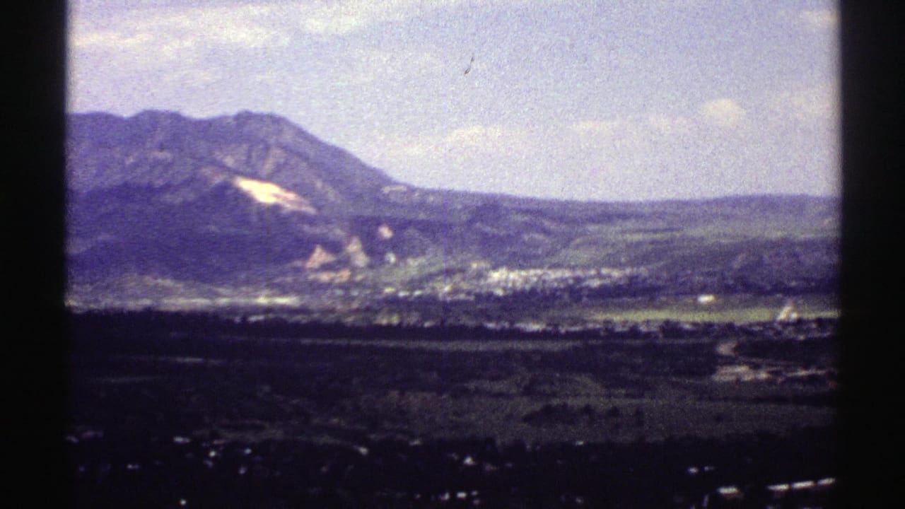 1969 Colorado 8mm frame showing mountains, a pale slope patch, and a distant valley settlement under a bright sky.