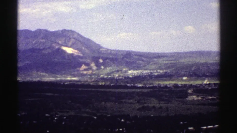 1969 Colorado 8mm frame showing mountains, a pale slope patch, and a distant valley settlement under a bright sky.