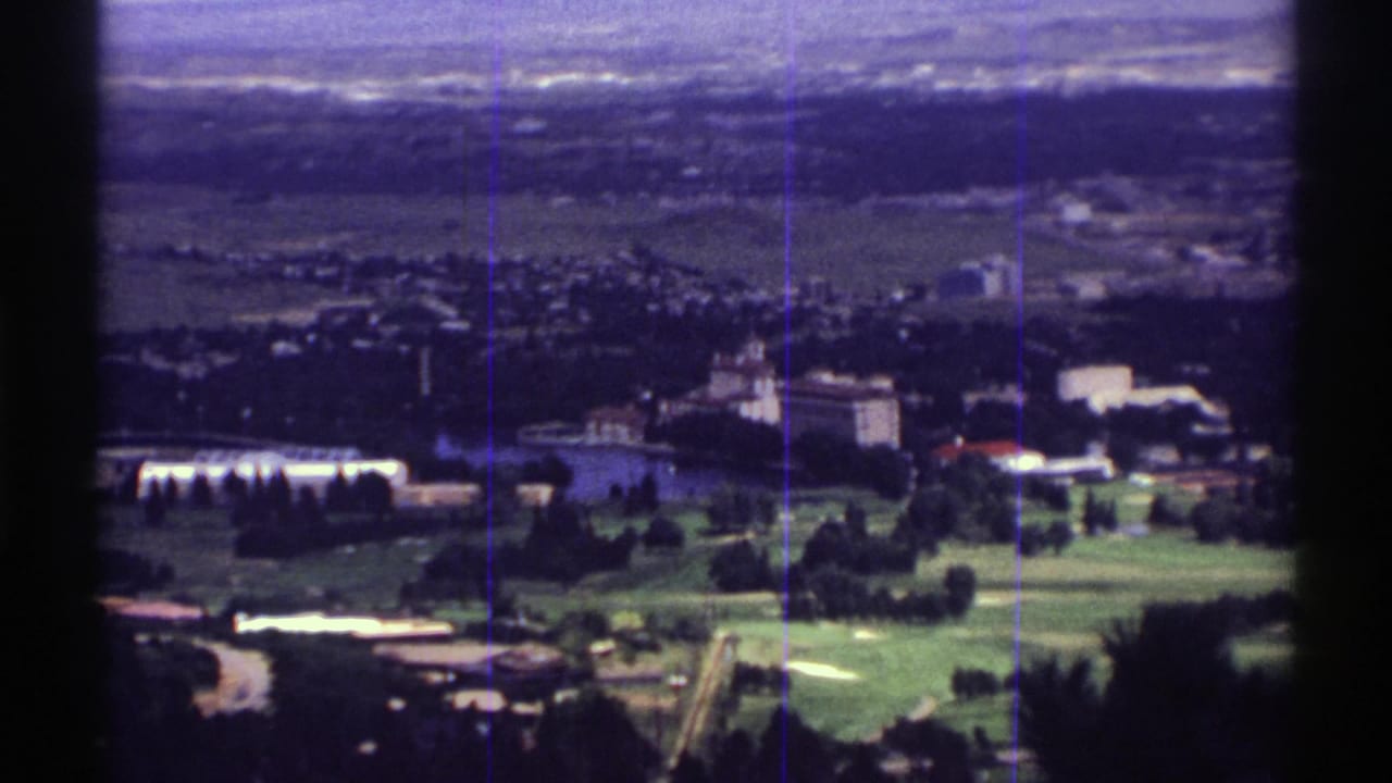 1969 Colorado home-movie frame showing green fields, clusters of buildings and distant terrain from a moving aerial or high vantage point.