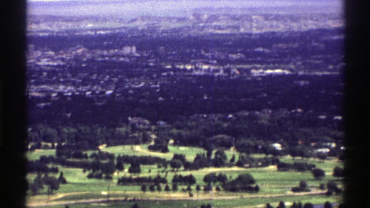 1969 Colorado aerial home-movie frame with fairways, trees, hillside homes, and a hazy built-up distance.