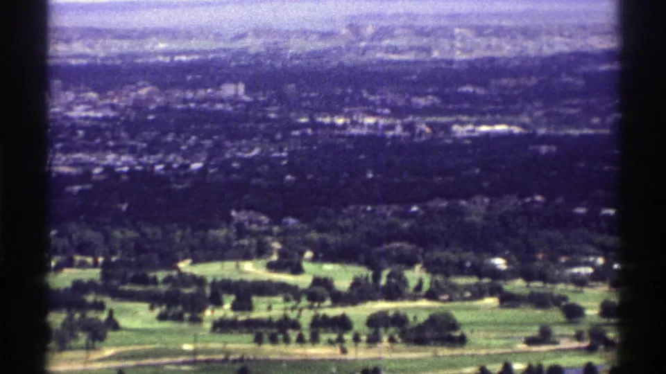 1969 Colorado aerial home-movie frame with fairways, trees, hillside homes, and a hazy built-up distance.