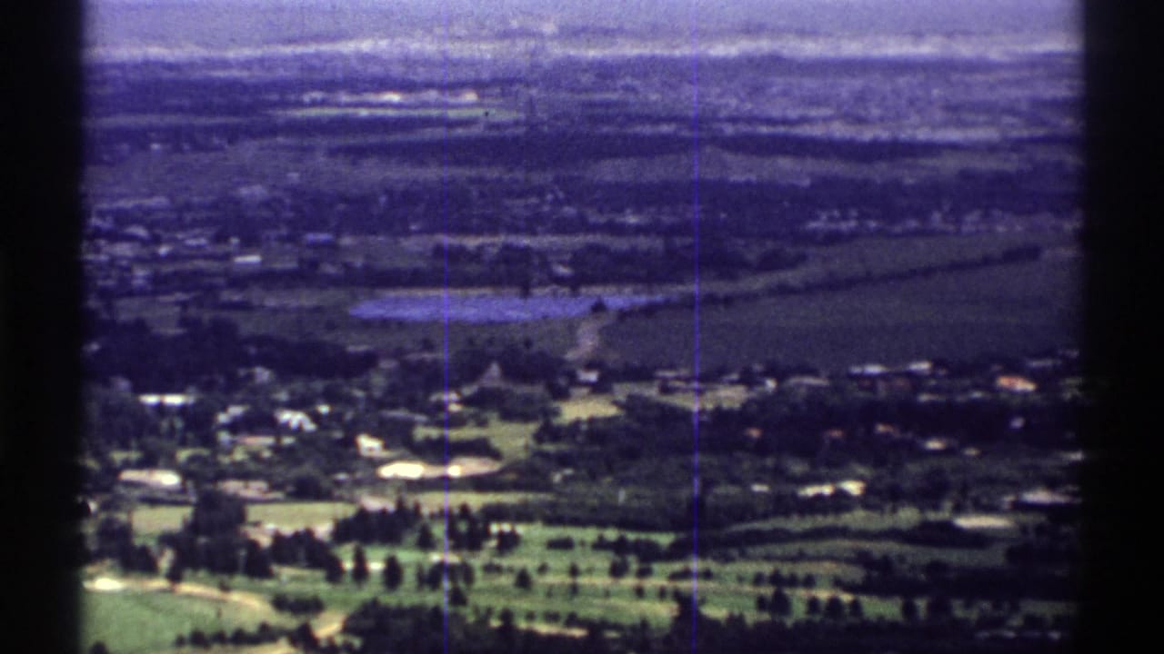 1969 Colorado home-movie frame with green fairways in front, low buildings and tree lines across a distant town landscape.