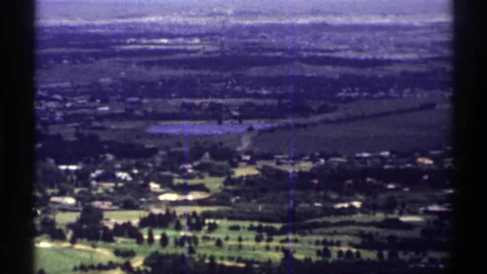 1969 Colorado home-movie frame with green fairways in front, low buildings and tree lines across a distant town landscape.