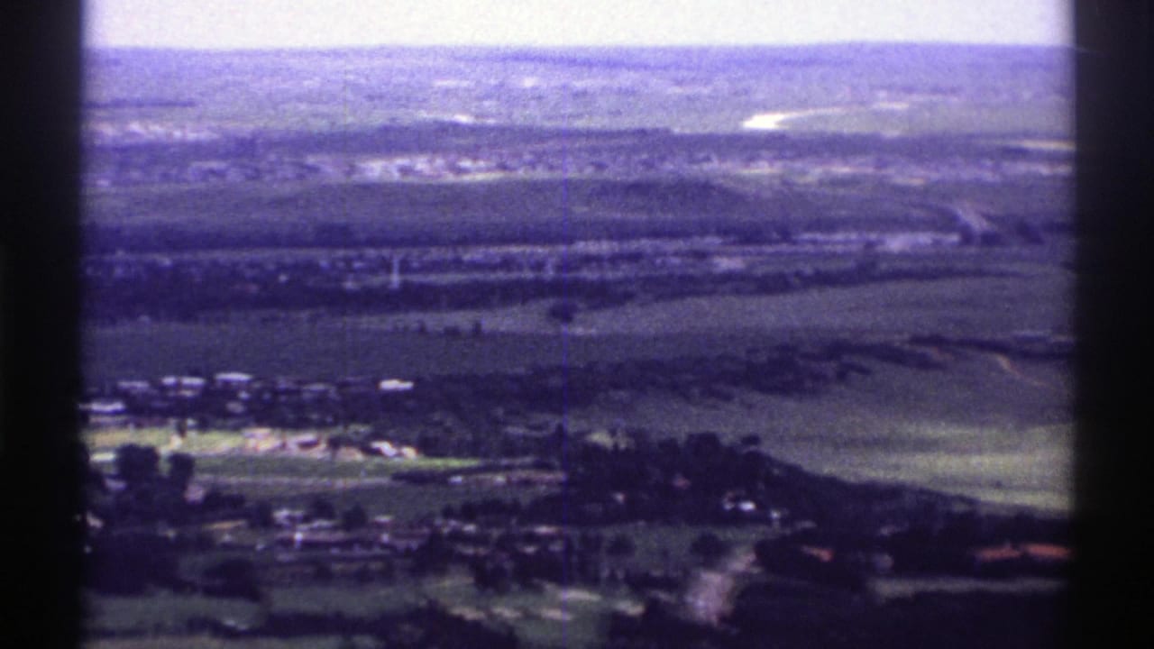 Aerial landscape with fields, small buildings, tree clusters, and distant low hills under a pale sky.