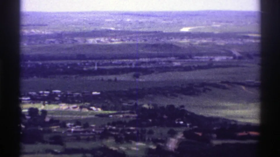 1969 Colorado airplane-view frame with green fields, scattered rural structures, tree clusters, and distant low hills softened by 8mm grain.