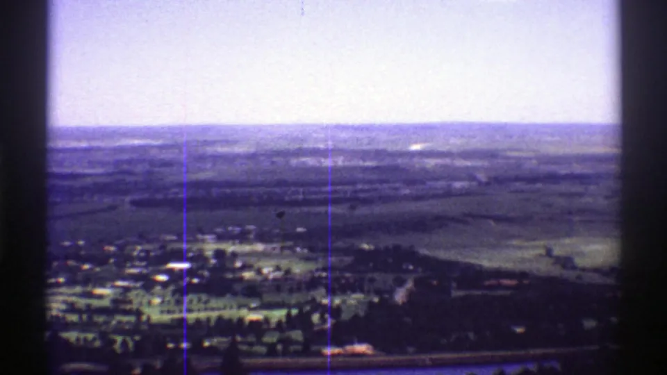1969 Colorado home-movie aerial frame showing a rural settlement edge, roads, open fields, and a wide horizon through soft 8mm color.