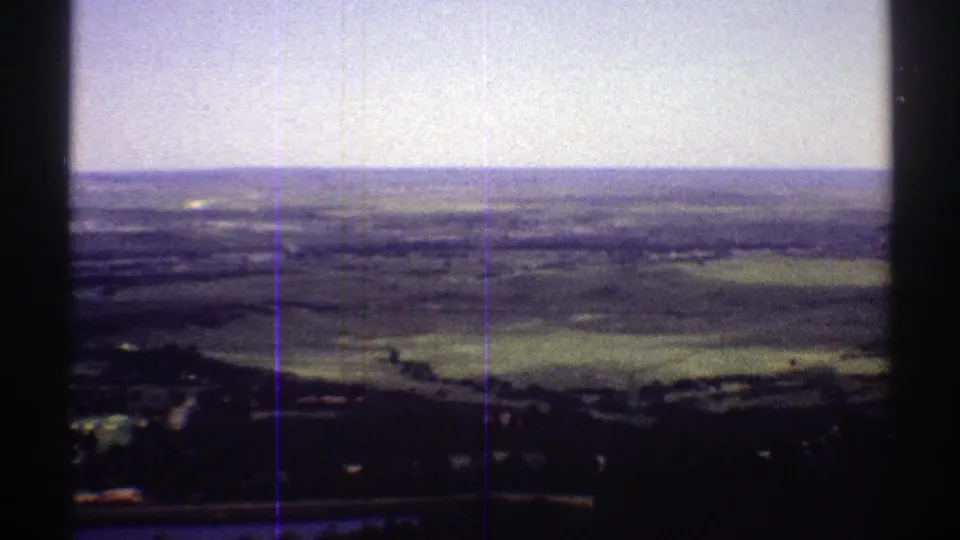 1969 Colorado 8mm view from an elevated overlook across fields, tree-dark slopes, and distant hills under a pale sky.