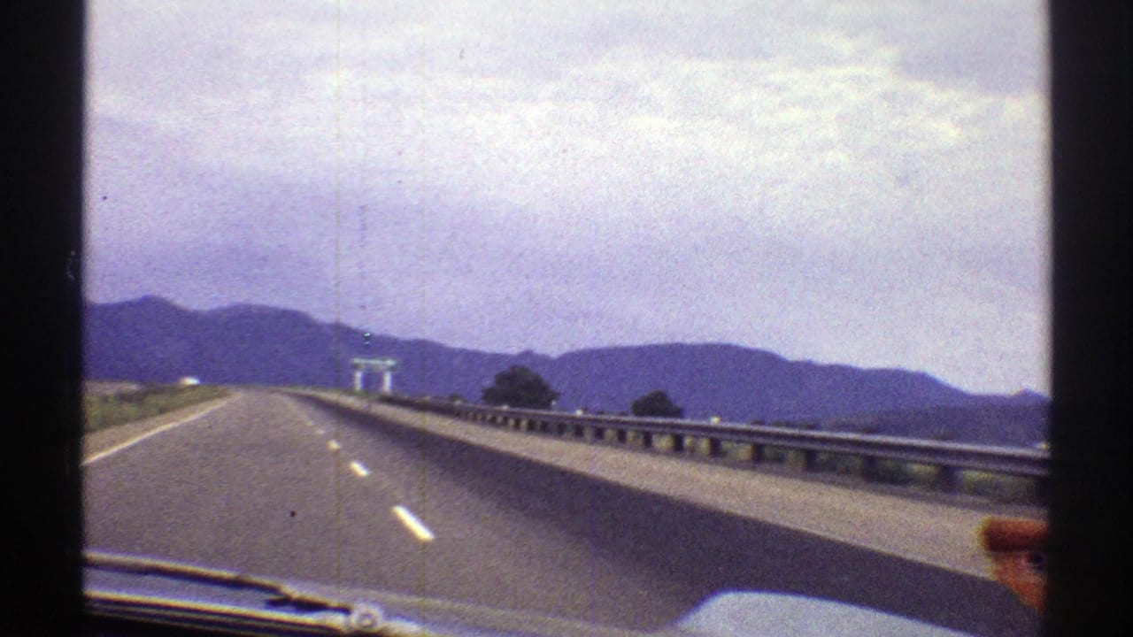 Highway seen from inside a moving car, with lane markings, guardrail, mountains, and cloudy sky.