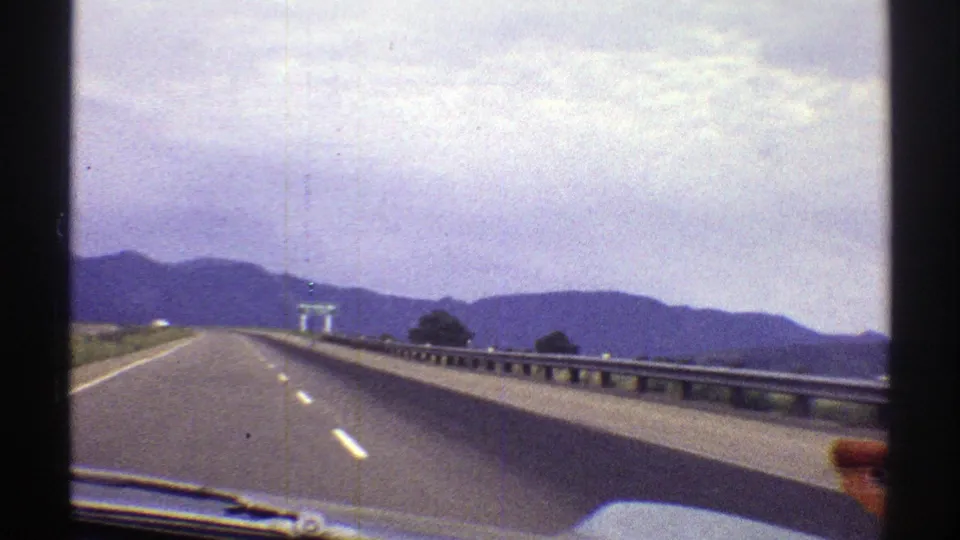 1969 Colorado 8mm view from a moving car showing an open highway, metal guardrail, distant mountains, and a bright cloudy sky.