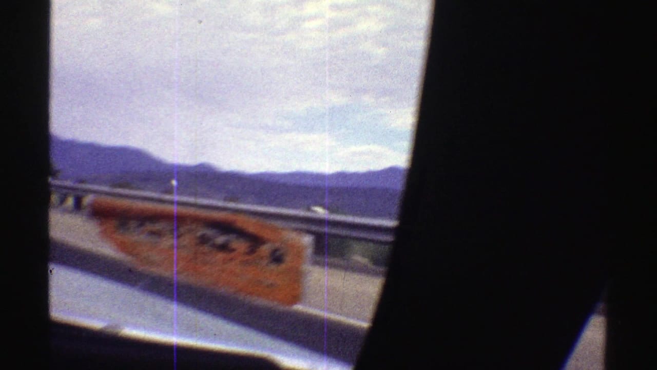 Car window view with a blurred orange roadside sign, guardrail, mountains, and dark interior frame.