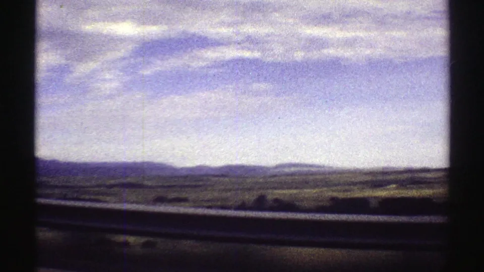 1969 Colorado road-trip view from inside a moving car, with guardrail, open land, distant mountains, and a wide partly cloudy sky.