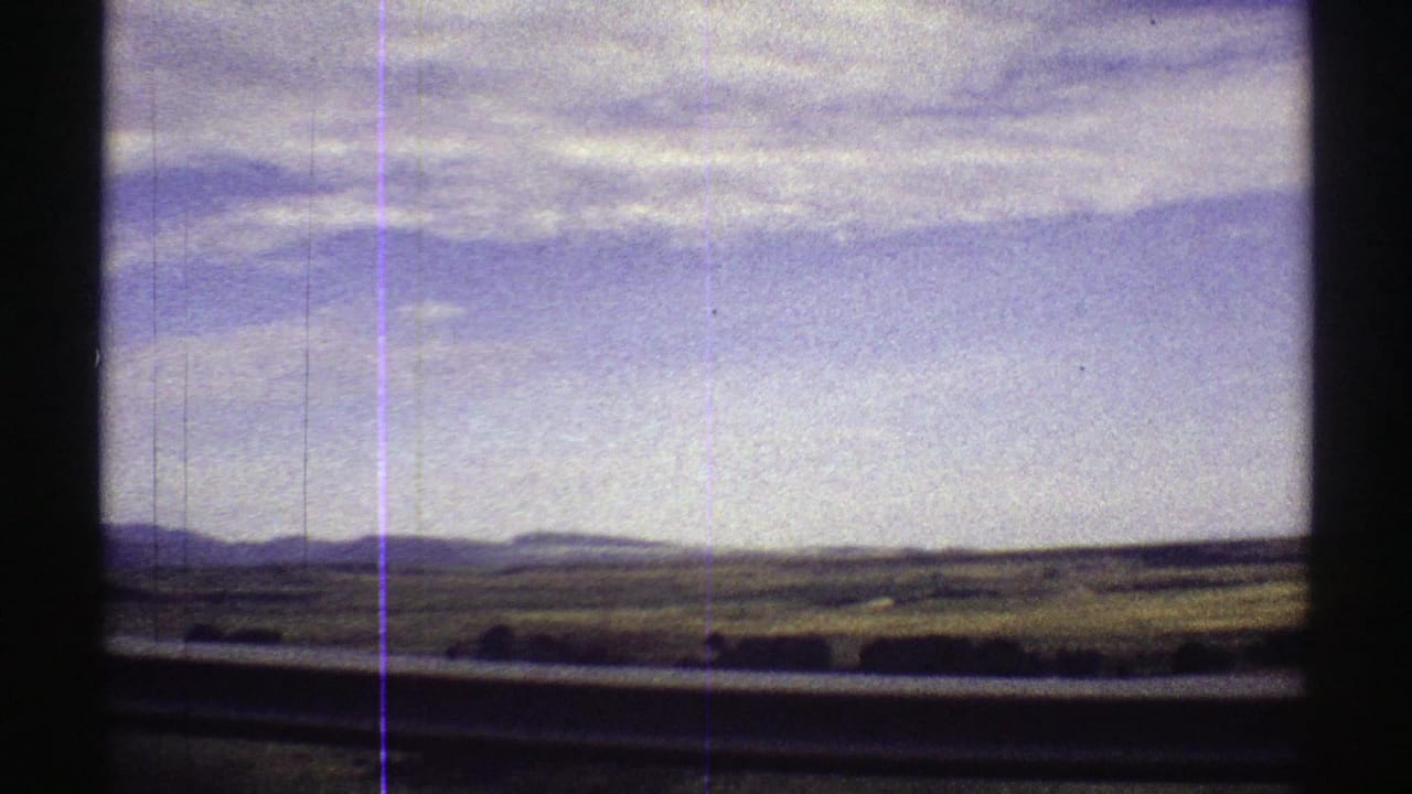 Green fields and distant mountains seen through dark vehicle framing under a cloudy blue sky.