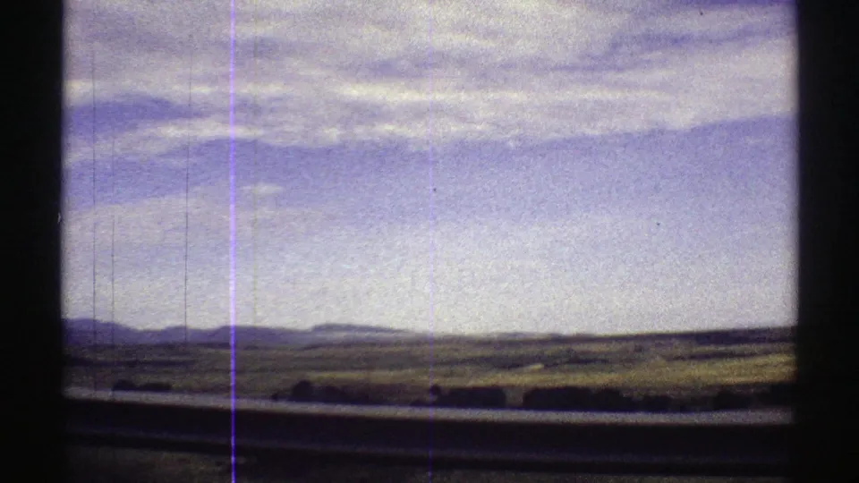 1969 Colorado plains viewed from a moving vehicle, with fields, distant mountains, and clouded sky partly blocked by dark frame edges.