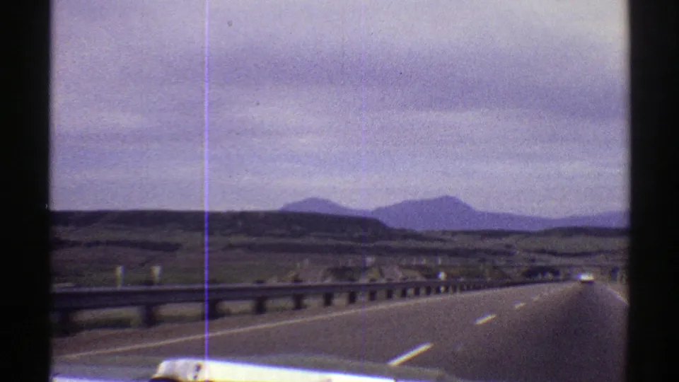 1969 Colorado road view showing a curving highway, guardrail, open plains, and a distant mountain horizon under a cloudy sky.