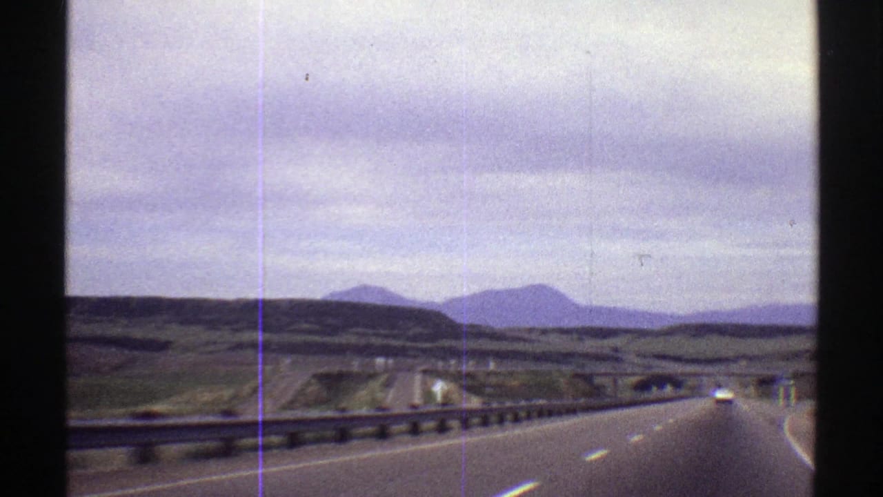 A moving-vehicle view on a Colorado highway in 1969, with road lanes, guardrails, distant cars, open hills, and low mountains visible through soft 8mm grain.