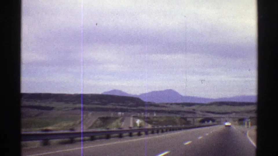 A moving-vehicle view on a Colorado highway in 1969, with road lanes, guardrails, distant cars, open hills, and low mountains visible through soft 8mm grain.