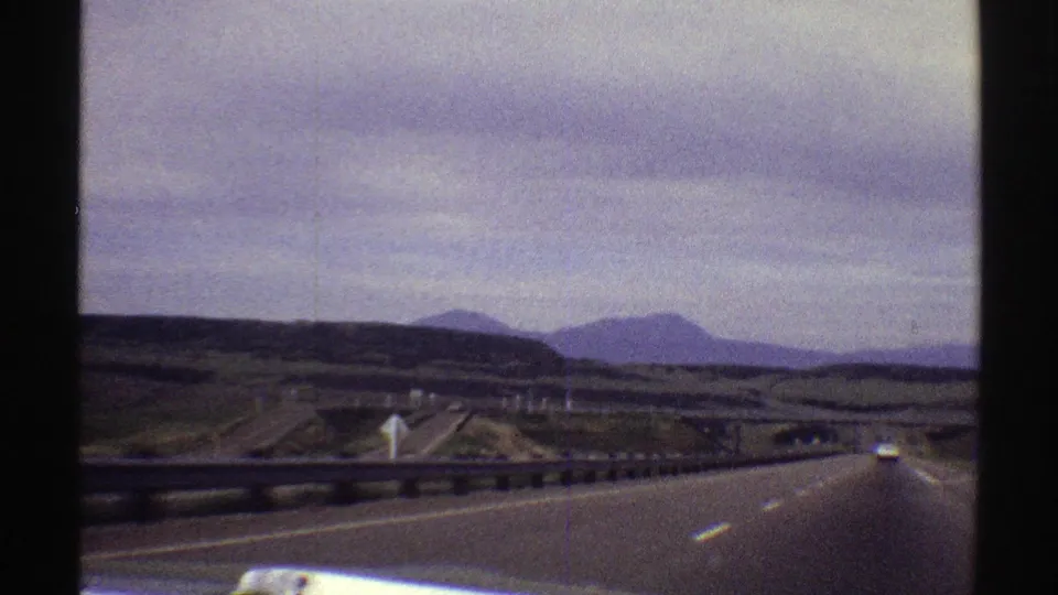 1969 Colorado home-movie frame from a vehicle, showing a highway, roadside guardrail, divided ramps or lanes, low hills, and muted mountains under an overcast sky.