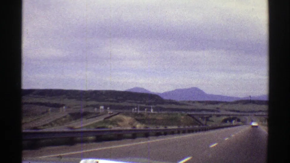 Final 1969 Colorado road-trip still showing a forward highway view, metal guardrail, sparse traffic, rolling terrain, and distant mountains in soft vintage 8mm color.