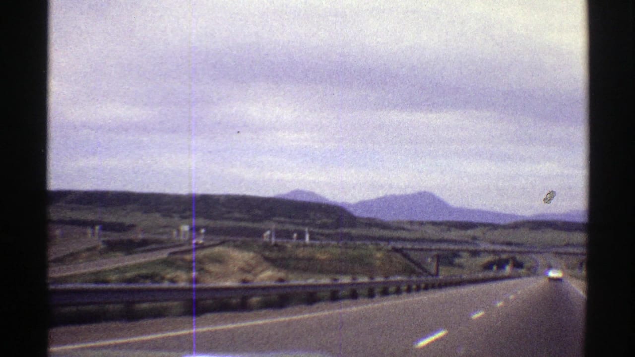 1969 Colorado 8mm road-trip view from a moving vehicle, looking along a highway toward a car ahead with rolling land and mountains in the distance.