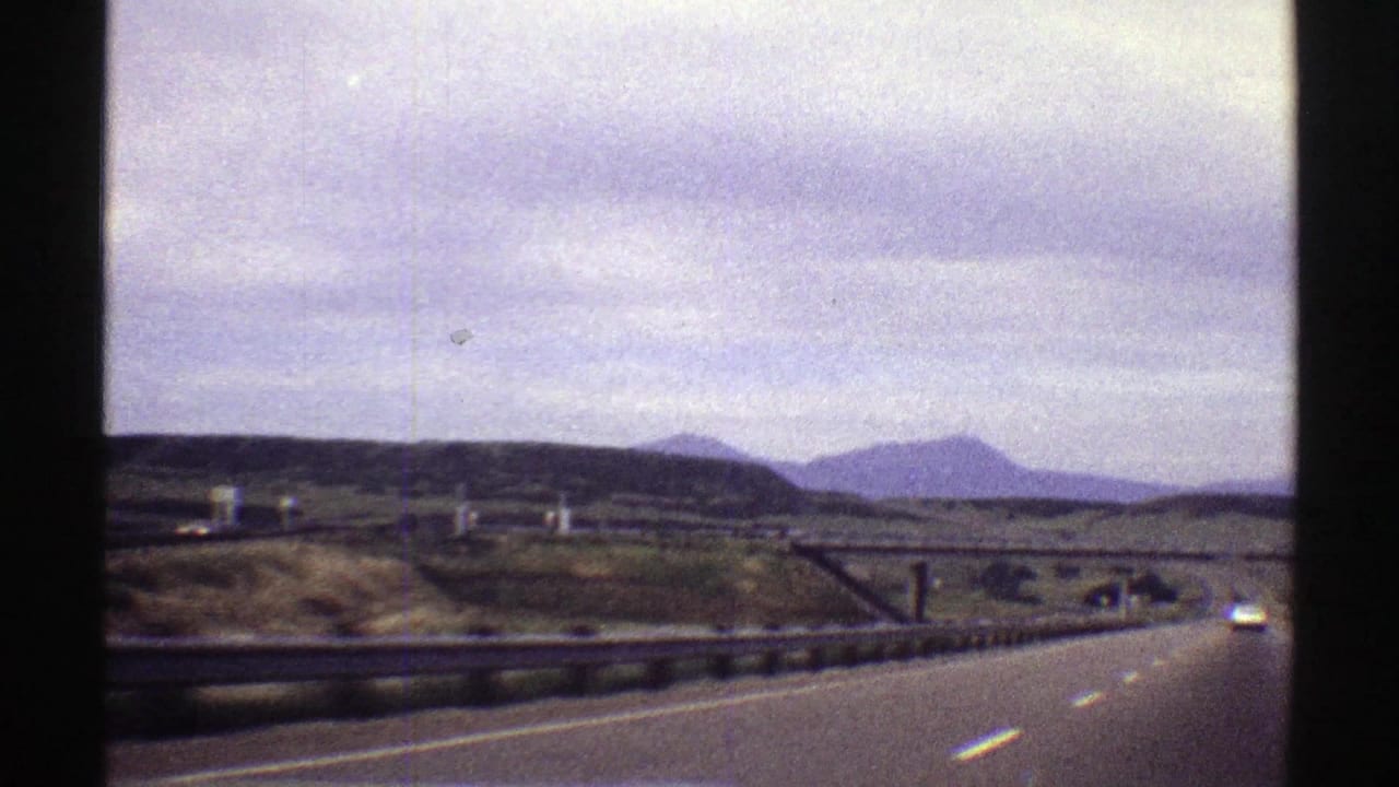 Highway seen from a moving vehicle with a car ahead, roadside barriers, fields, and mountains on the horizon.
