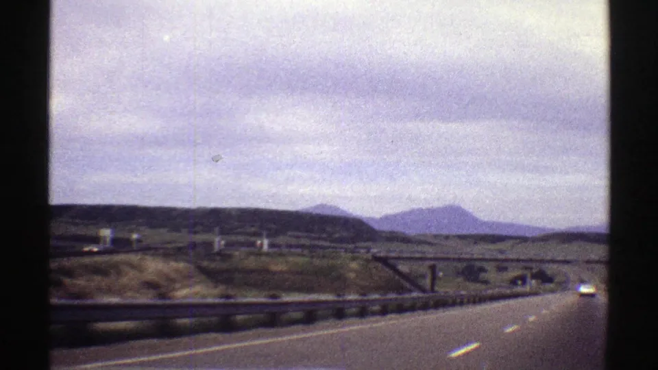 Forward-facing 1969 Colorado highway footage shows a car ahead on the road, low guardrails, open fields, and hazy mountain shapes beneath a cloudy sky.