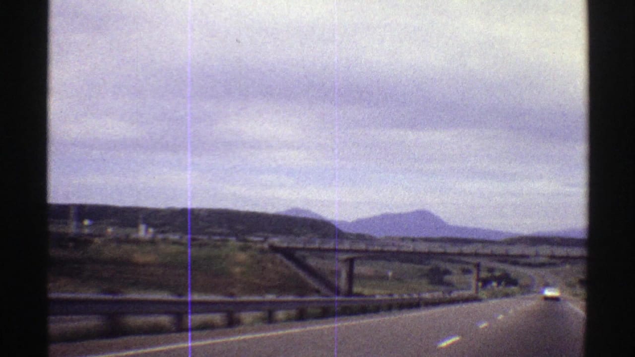 1969 Colorado road-trip frame from inside a vehicle, approaching a concrete overpass while a car travels ahead and mountains sit beyond the highway.