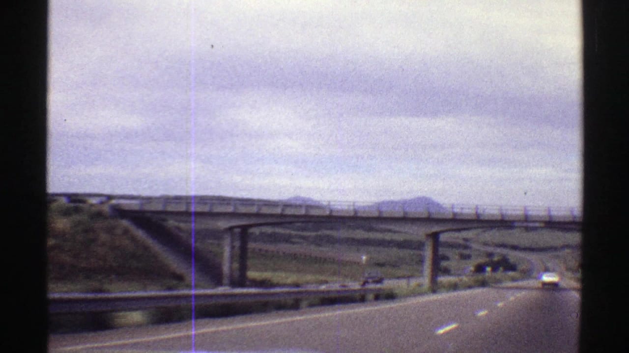 Late-1960s Colorado 8mm frame shows a highway scene beside a concrete overpass, with a car ahead, guardrail, open roadside, and mountain horizon.
