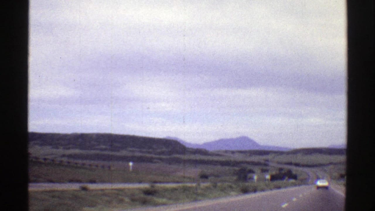 1969 Colorado 8mm driving shot with a car ahead, open road, rolling hills, and muted mountain forms.