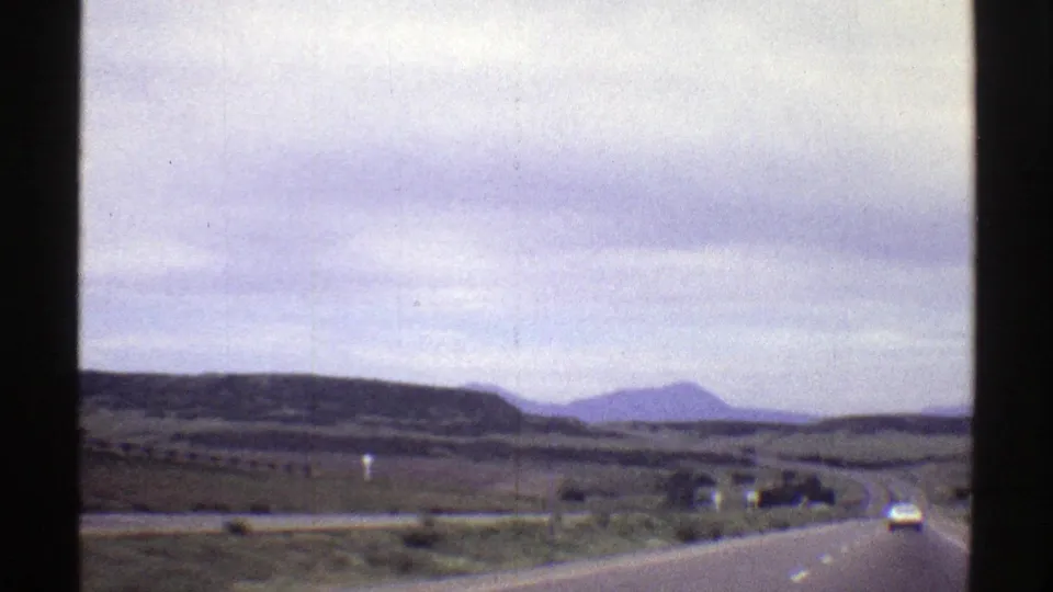 1969 Colorado 8mm driving shot with a car ahead, open road, rolling hills, and muted mountain forms.